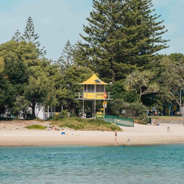 vibrant yellow lifeguard tower nestled among tall trees on the beach, viewed from the water, representing a typical and picturesque scene for those travelling alone in the Gold Coast