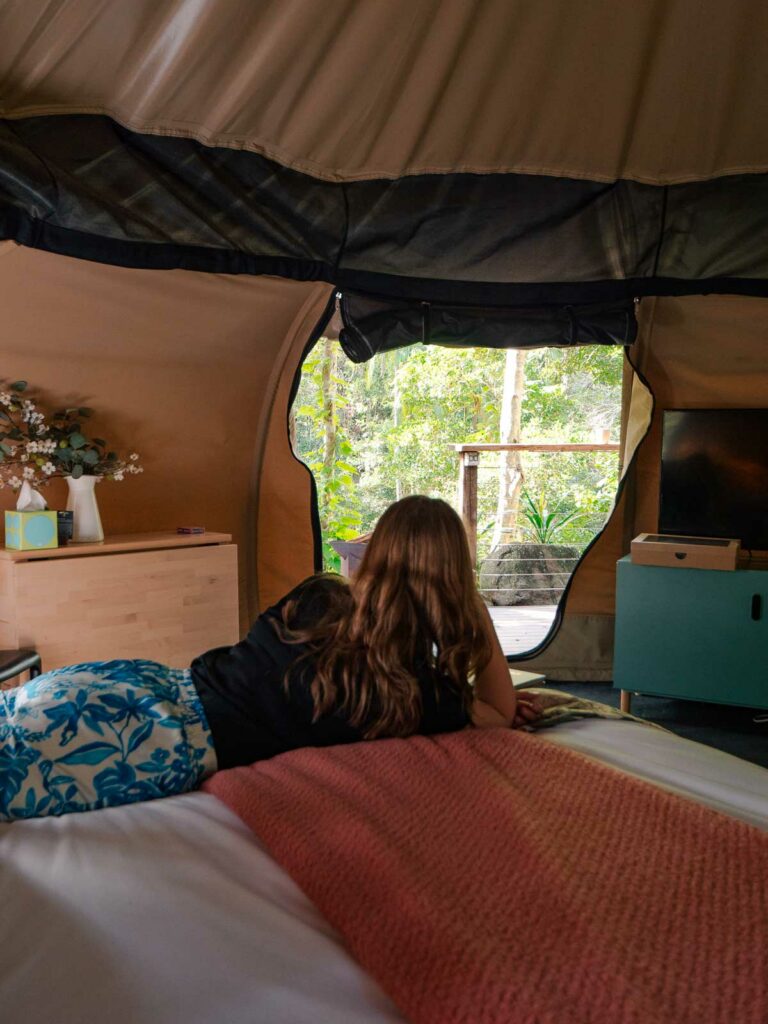 A woman relaxes on a comfortable bed inside a glamping tent, gazing out at the tranquil forest scenery through the open tent flap, blending luxury and nature