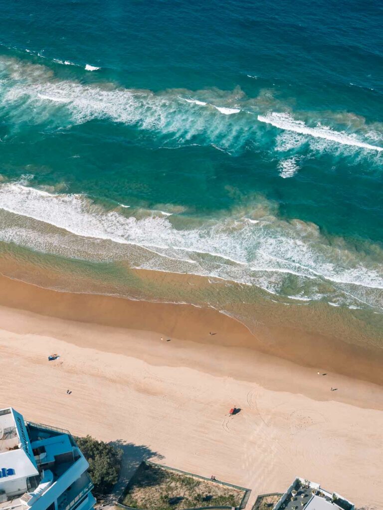 An aerial view of the beach shows waves crashing onto the sandy shore, with a few people enjoying the coastline.