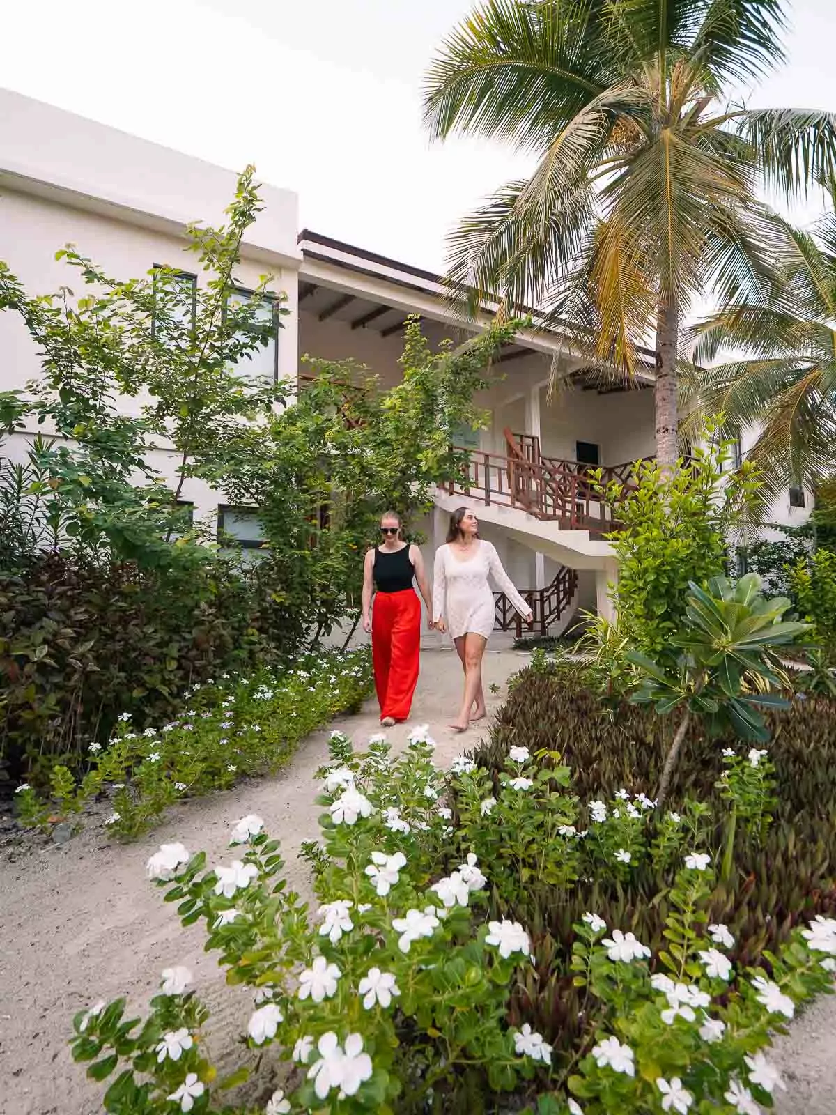 Two women walking on a sandy path surrounded by lush tropical greenery and white flowers, leading to a two-story building at OBLU XPERIENCE Ailafushi