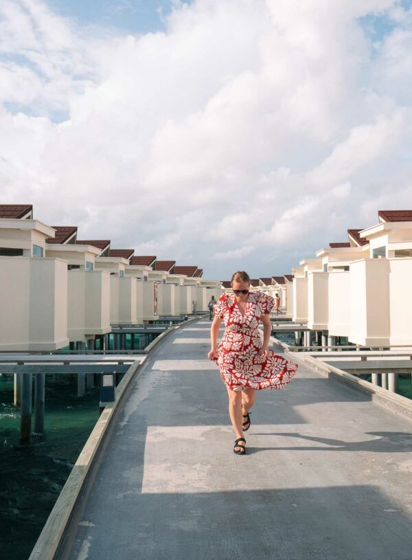 A woman walking along the path between overwater villas at OBLU XPERIENCE Ailafushi, a budget Maldives resort, with a cloudy sky overhead
