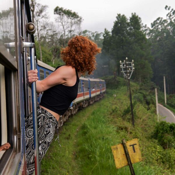 A woman with curly hair leans out of a moving train, enjoying the scenic views while travelling in Sri Lanka