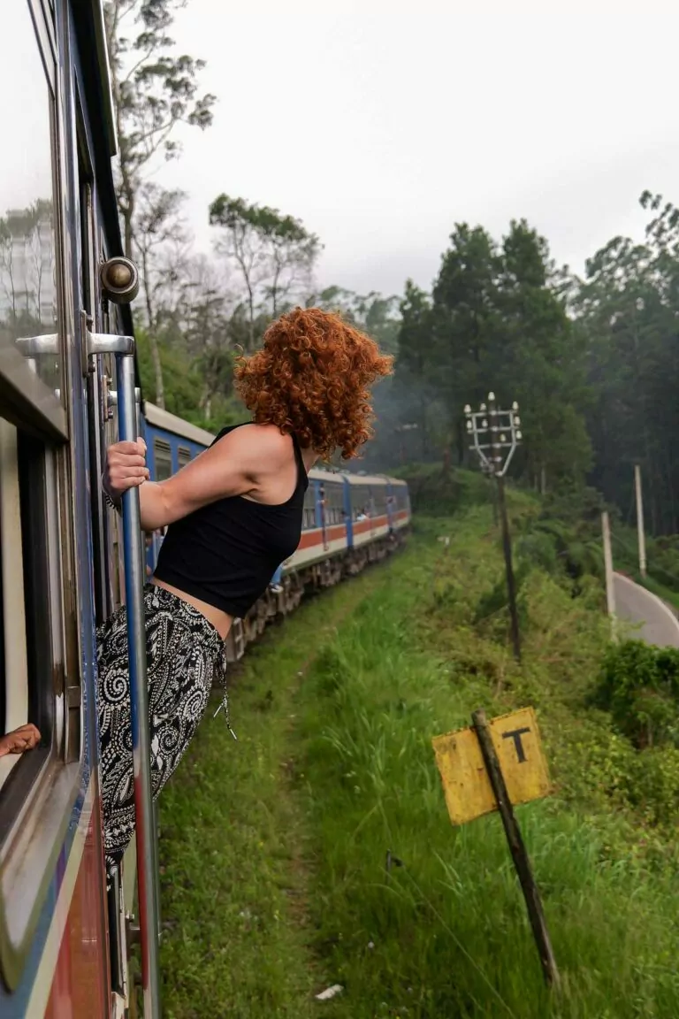 A woman with curly hair leans out of a moving train, enjoying the scenic views while travelling in Sri Lanka