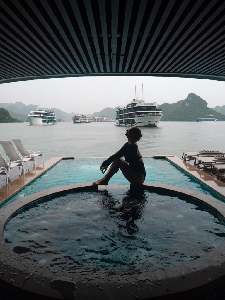 A woman in a black swimsuit sitting at the edge of a jacuzzi on the Capella Cruise, with several cruise boats in the background on Halong Bay.