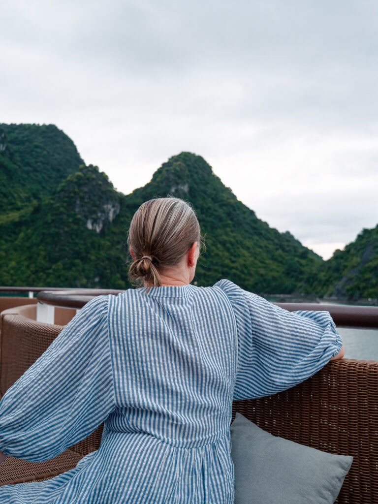 A woman relaxing on a deck chair aboard the Capella Cruise, gazing at the towering green limestone islands of Lan Ha Bay.