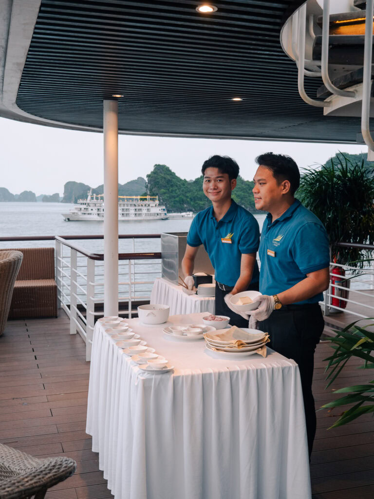 Two Capella Cruise staff members setting up a table on the deck, with Halong Bay’s limestone islands visible in the background.