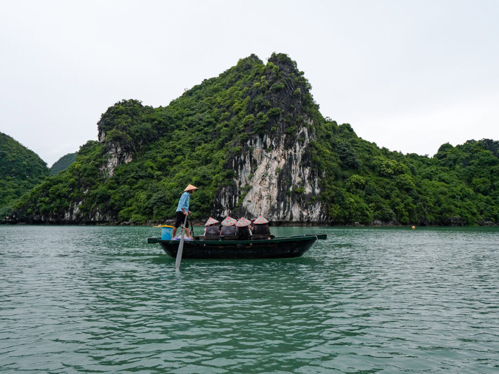 A person rowing a small boat past limestone cliffs in Halong Bay, with traditional woven hats arranged neatly behind them.