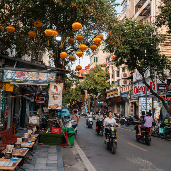 A busy street in Hanoi lined with shops, restaurants, and motorbikes, decorated with hanging lanterns, showcasing a typical scene from a Hanoi itinerary.