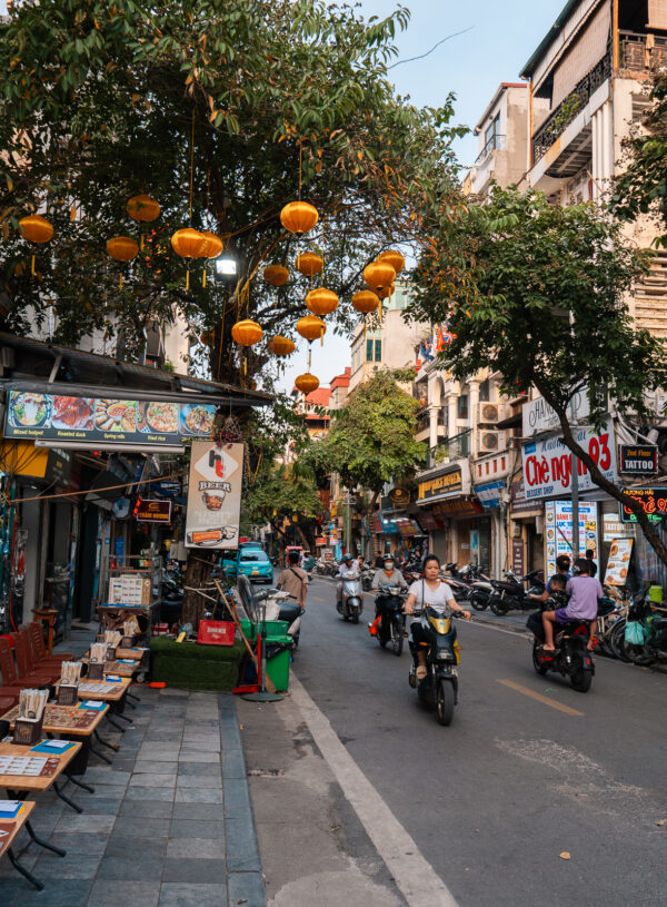 A busy street in Hanoi lined with shops, restaurants, and motorbikes, decorated with hanging lanterns, showcasing a typical scene from a Hanoi itinerary.