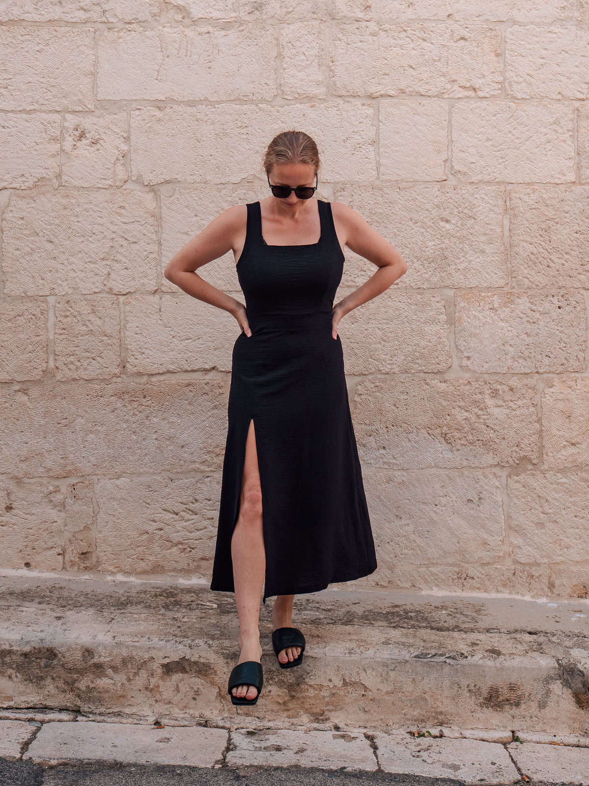 A woman in a sleek, sleeveless black midi dress with a high slit from Milan Linen tailor in Hoi An, paired with black slide sandals, standing in front of a neutral stone wall