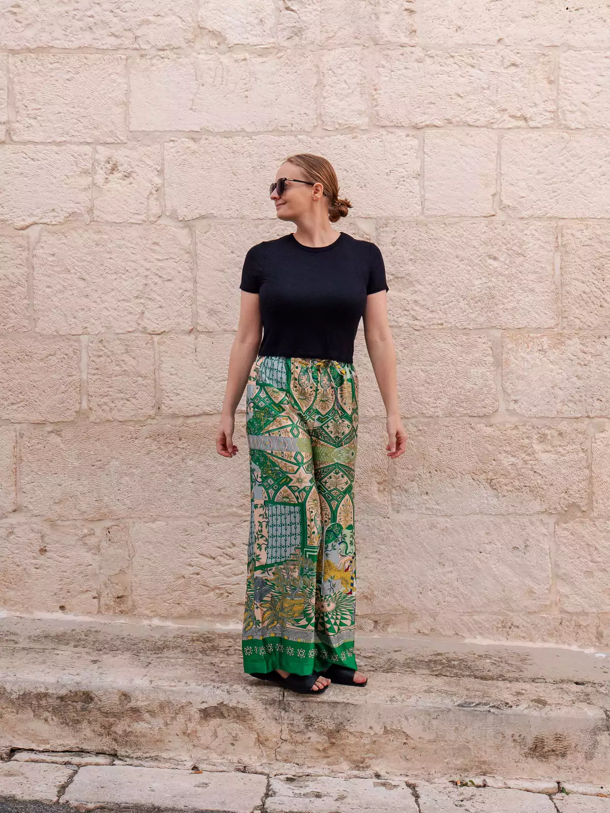 A woman in green and blue patterned wide-leg pants custom tailored at Yaly Couture and a black fitted t-shirt stands in front of a stone wall
