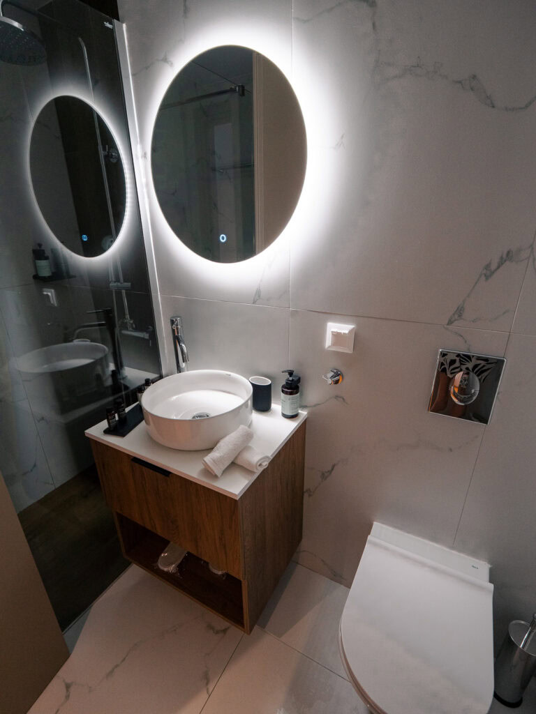 A modern bathroom with a round illuminated mirror above the sink and clean marble finishes.