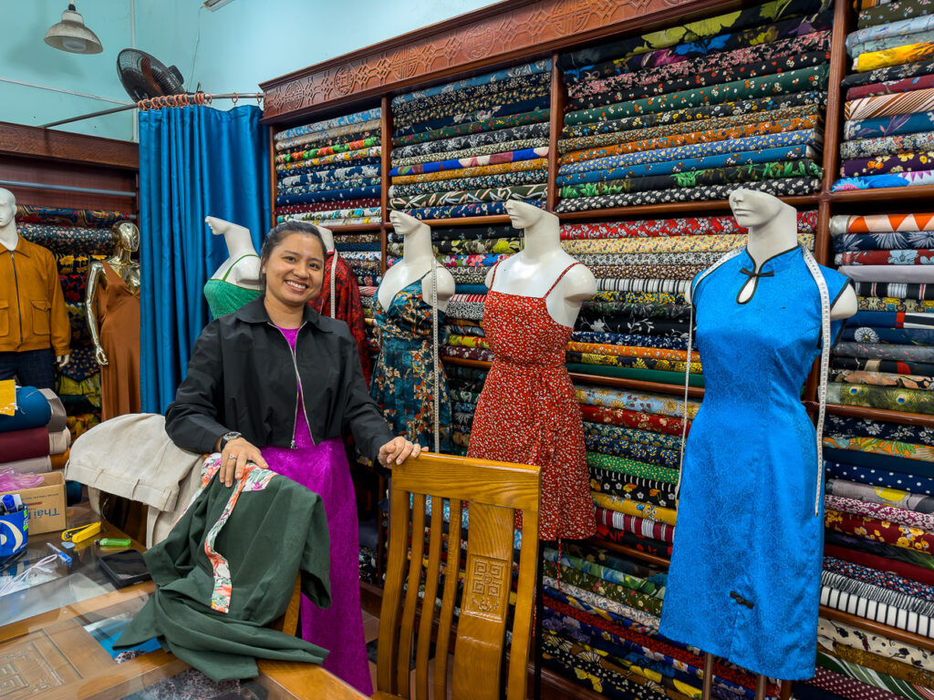 Ly at Pink Lotus tailor in Hoi An, beside the fabric display