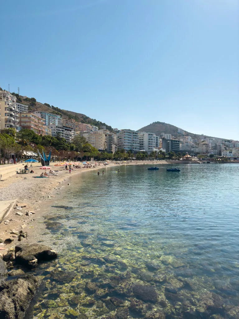 Solo travel in Albania: Saranda's curved beachfront with crystal-clear shallows revealing seabed, modern apartments rising up hillside.