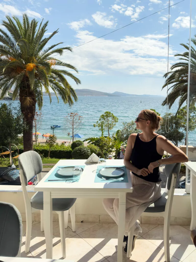 A solo female traveller dines at a seafront restaurant in Albania, with palm trees and sea views providing a Mediterranean backdrop.