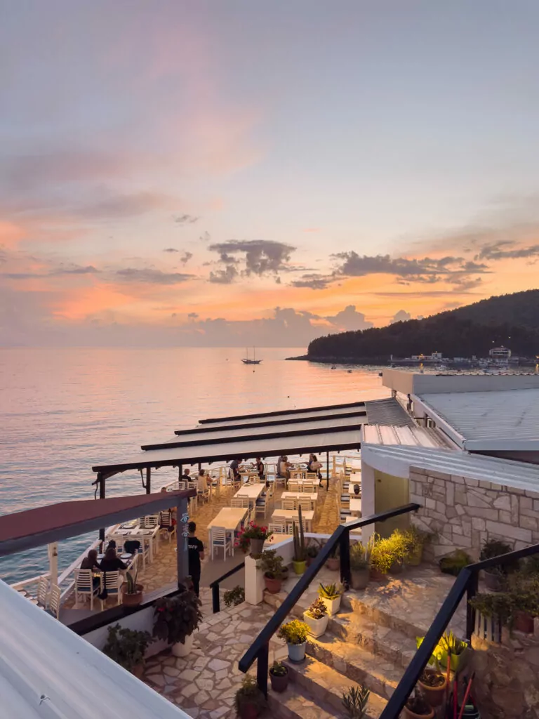 Rooftop bar at sunset in Himare, tiered seating overlooking golden-lit bay and mountains.