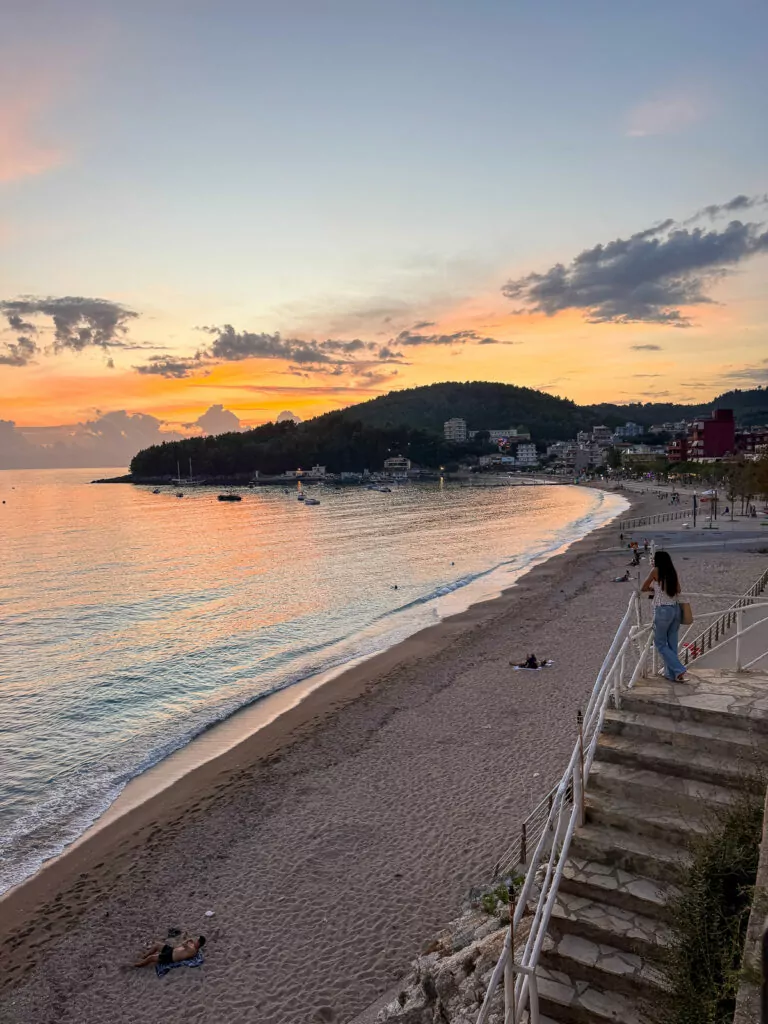 Female solo traveller walks along Himare sunset promenade, golden light illuminating curved coastline and steps.