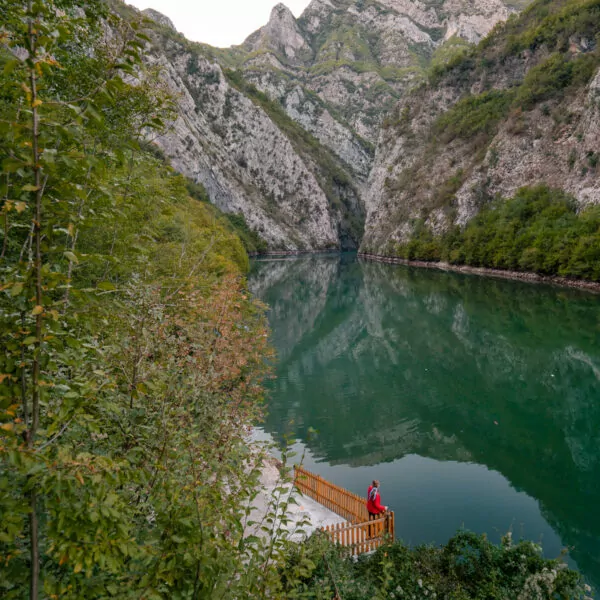 Solo traveller in red stands on viewing platform overlooking Lake Koman's dramatic gorge, emerald waters winding between limestone cliffs.