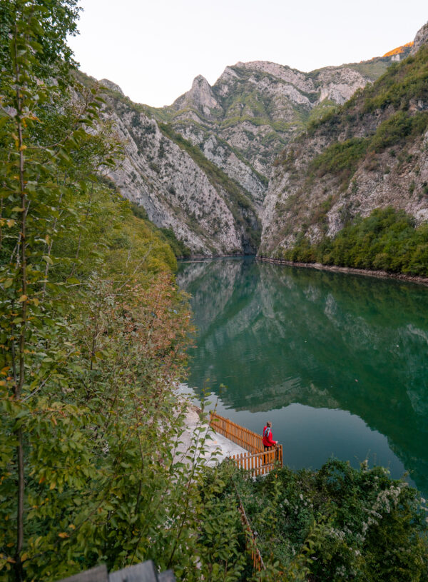 Solo traveller in red stands on viewing platform overlooking Lake Koman's dramatic gorge, emerald waters winding between limestone cliffs.