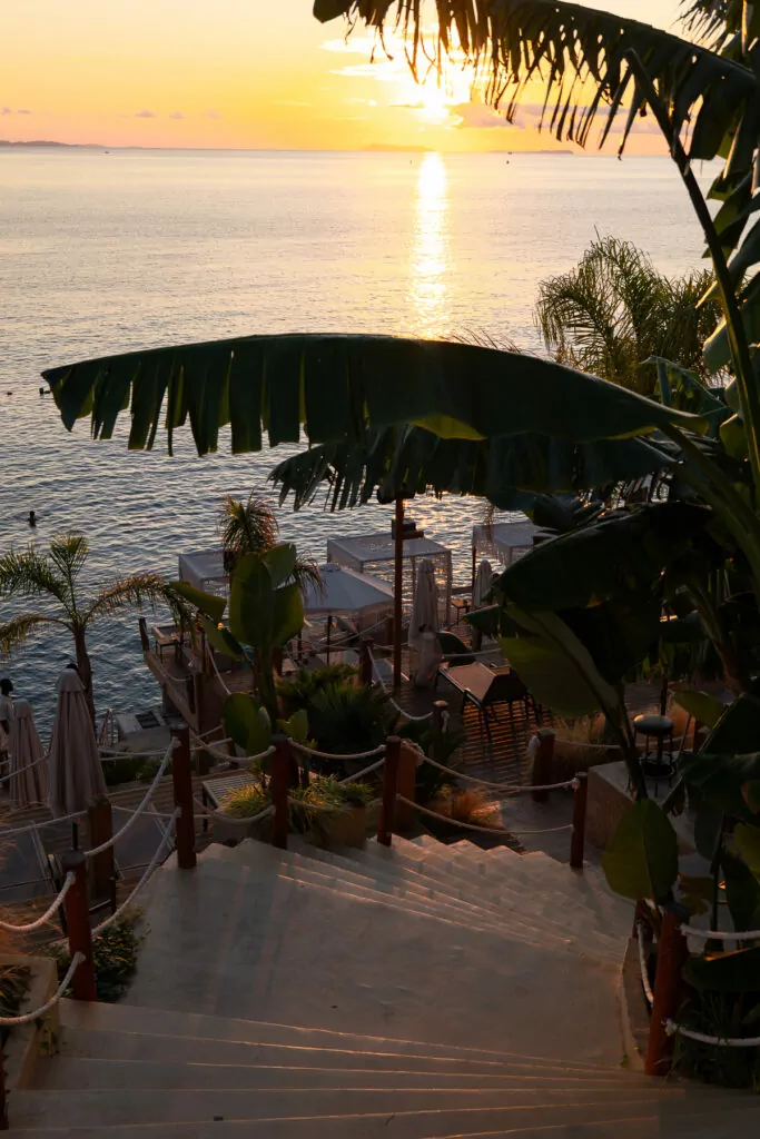 Palm-fringed steps lead down to a beach bar at sunset in Sarandë, creating an atmospheric coastal scene.