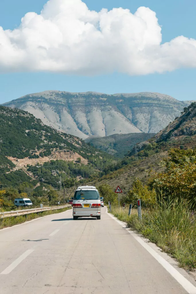 Solo road trip through Albania's mountains reveals winding roads and dramatic peaks, a white car providing scale against the vast limestone landscape.