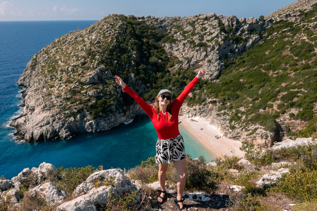 Female traveller in red jacket celebrates atop cliffs overlooking Grama Bay Beach's horseshoe bay, surrounded by Mediterranean waters.