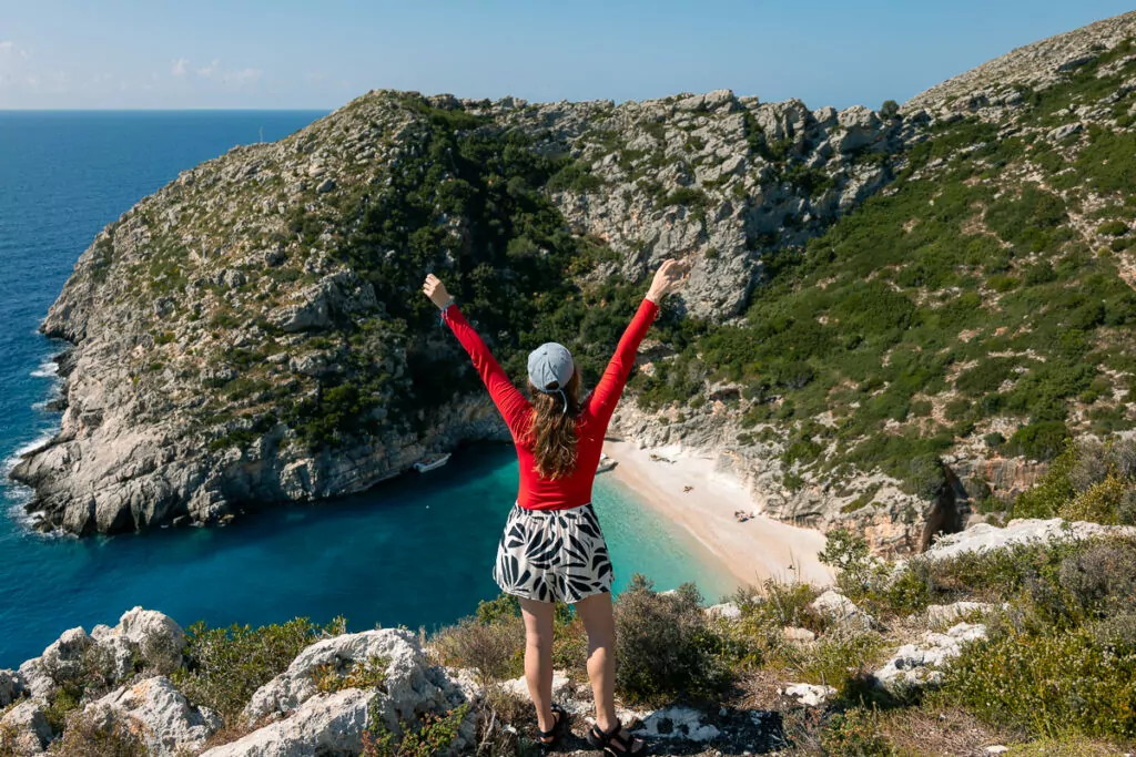 Person celebrates atop cliffs overlooking Grama Bay, Albania, with turquoise waters and limestone formations below