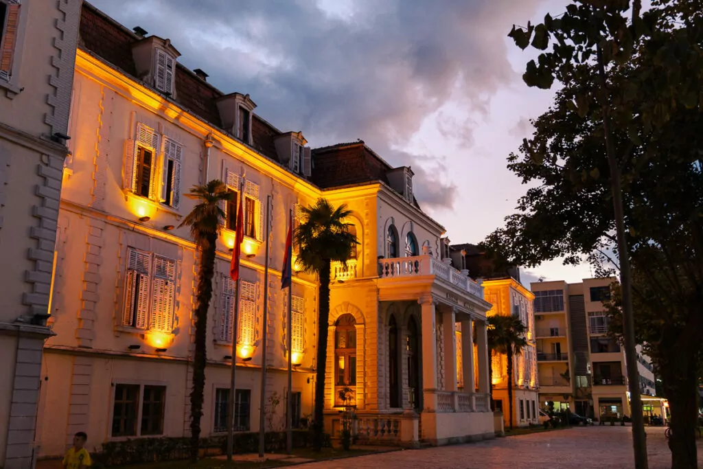 Italian-influenced architecture glows warmly at dusk in Shkodër, ornate yellow buildings illuminated by golden lights.