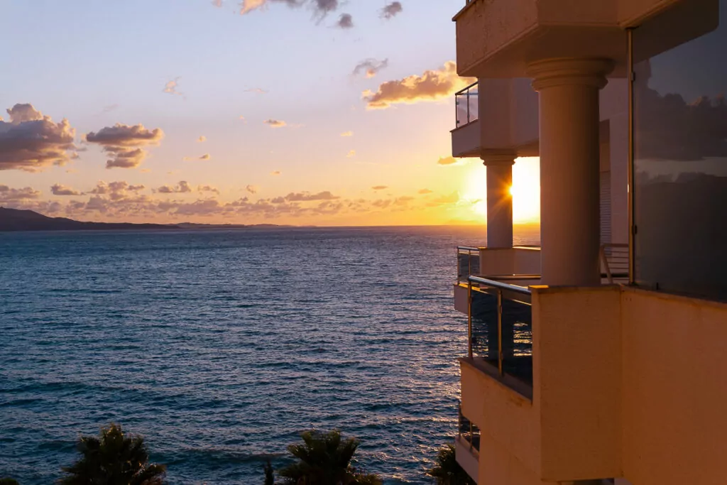 Warm orange sunset colours reflect off calm Mediterranean waters, viewed from a Sarandë apartment balcony with white concrete columns.