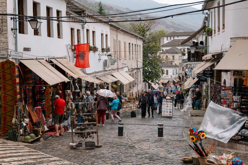 Bustling cobblestone street in Gjirokaster's bazaar where solo travellers browse traditional shops displaying red garments and souvenirs against white-walled buildings on a misty morning.