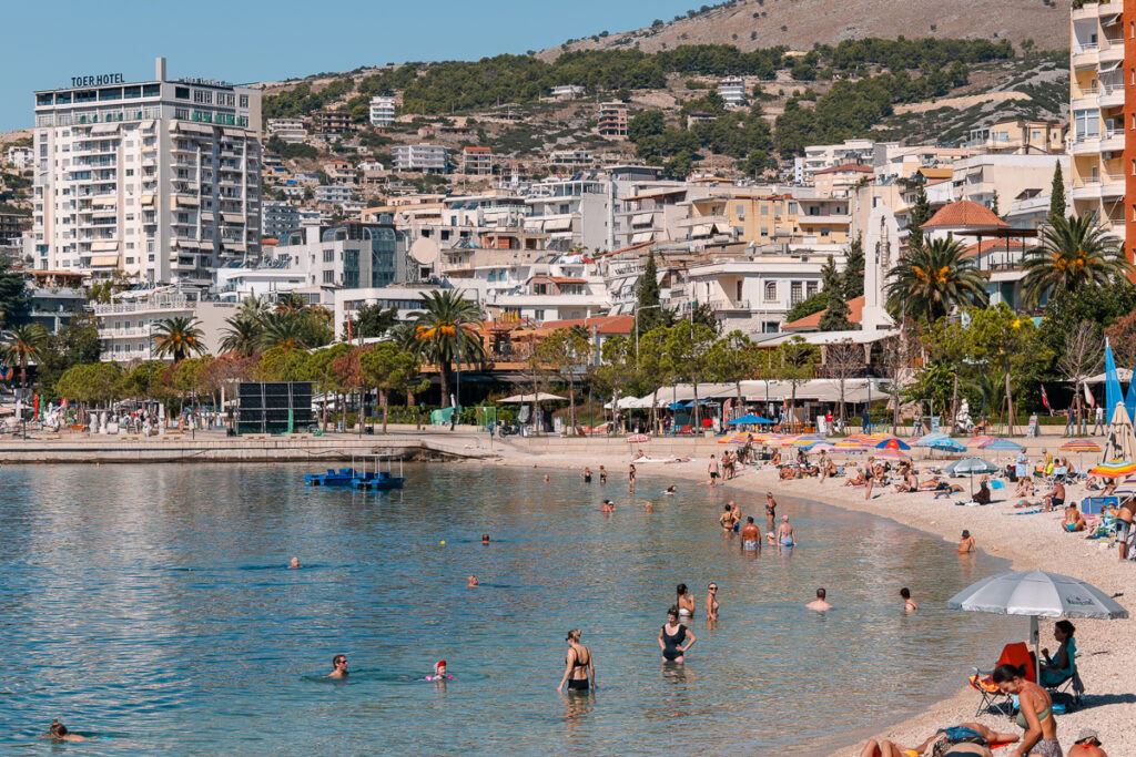 Beachgoers wade in Saranda's clear bay waters while apartments and hotels line the curved promenade, mountains rising behind the city.