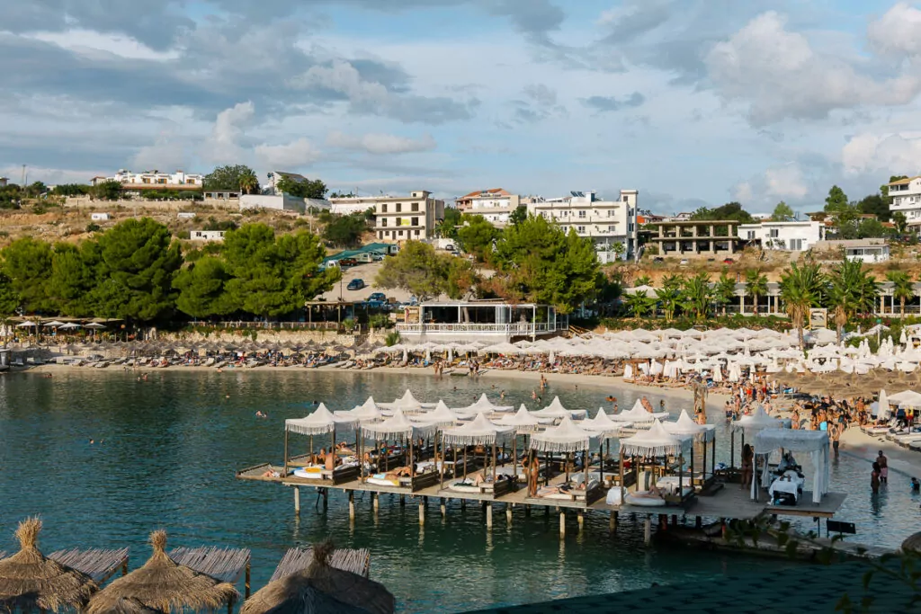 Ksamil beachfront restaurants with wooden pier structures extending over turquoise waters, buildings terraced up green hillside.