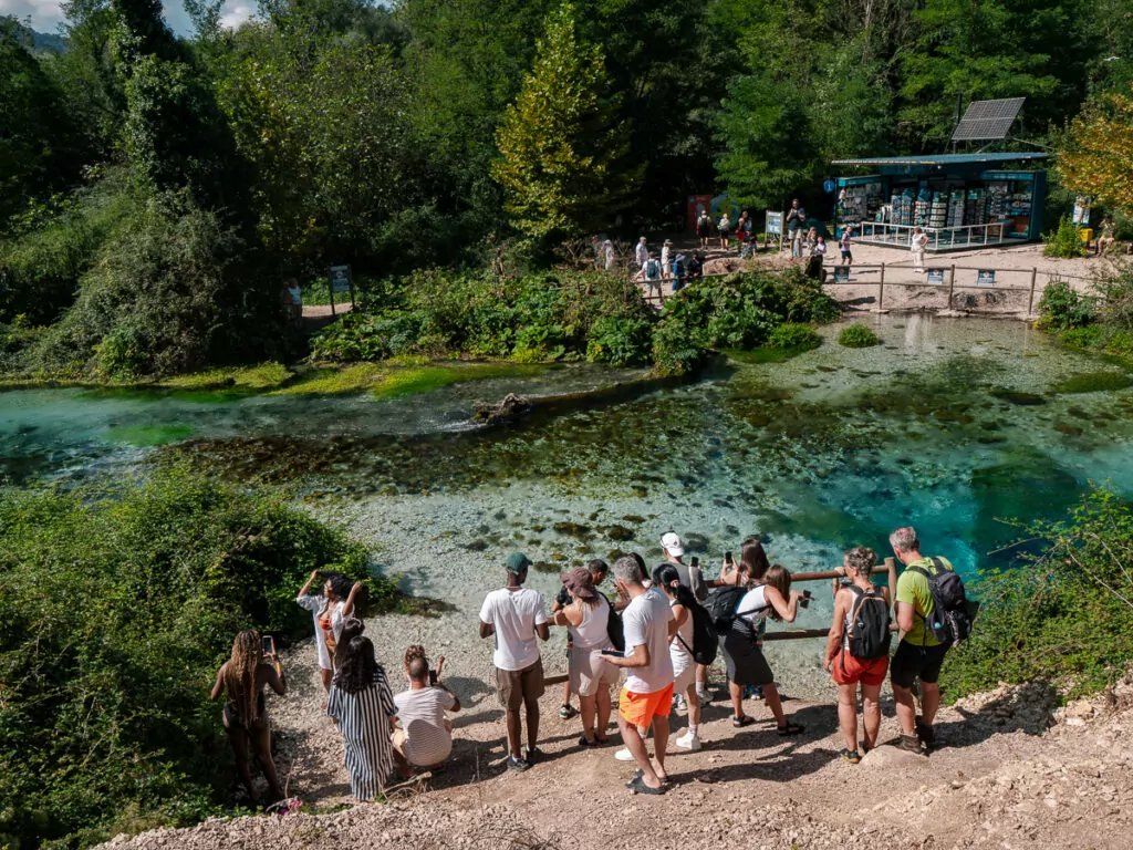 Tourists gather around Blue Eye Spring's vivid turquoise pool, surrounded by lush summer vegetation.