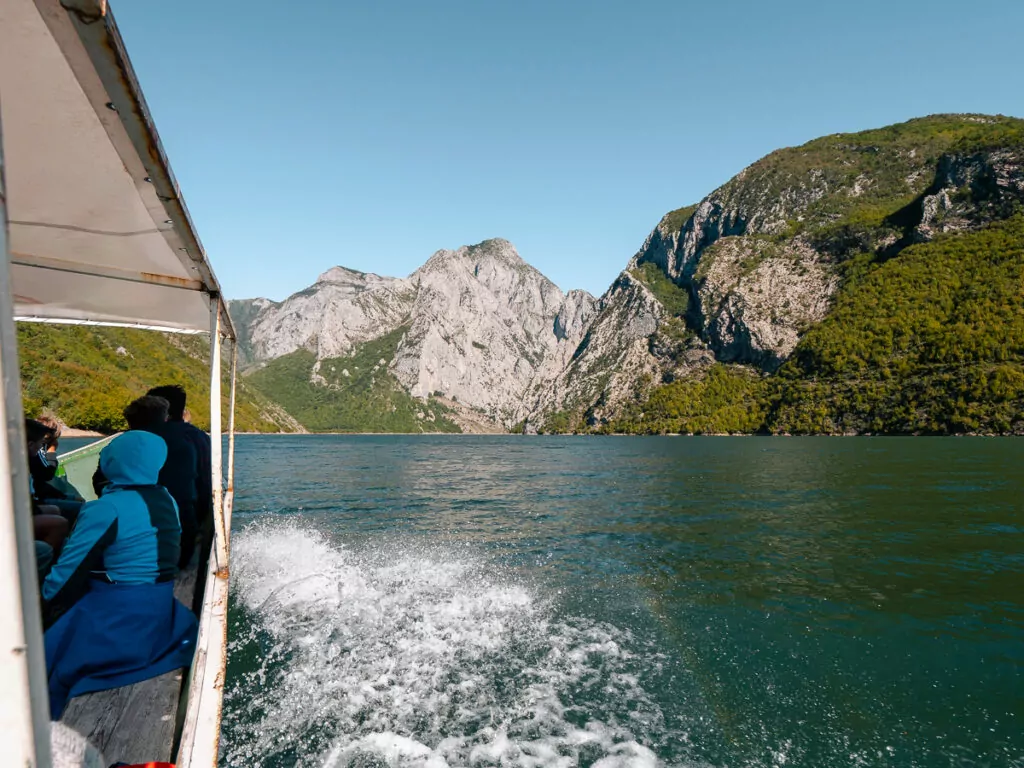 Water churns white wake behind Lake Koman ferry as solo adventurer in blue jacket gazes at vertical grey-brown cliffs plunging into deep green waters.