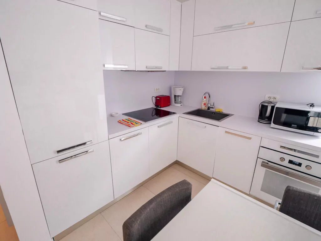 Modern white L-shaped kitchen in a rental apartment, featuring sleek cabinets and essential appliances with red accents.