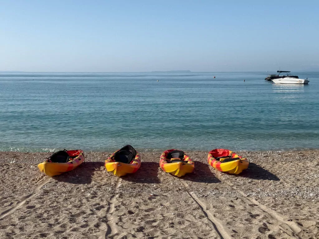 Four vibrant kayaks - yellow and orange - rest on pebbly Ksamil Beach, with turquoise waters stretching to a distant motorboat under clear blue skies.