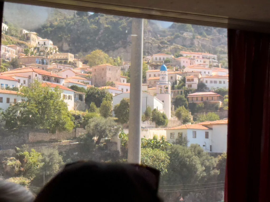 A traditional Albanian hillside village called Dhërmi, complete with white mosque minaret, sits nestled amongst terracotta rooftops viewed through an apartment window.