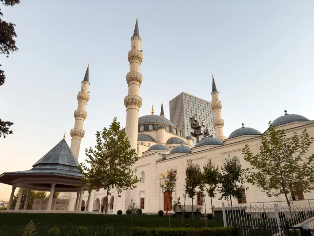 Et'hem Bey Mosque's elegant cream domes and minarets rise against clear blue Tirana sky, surrounded by manicured gardens.