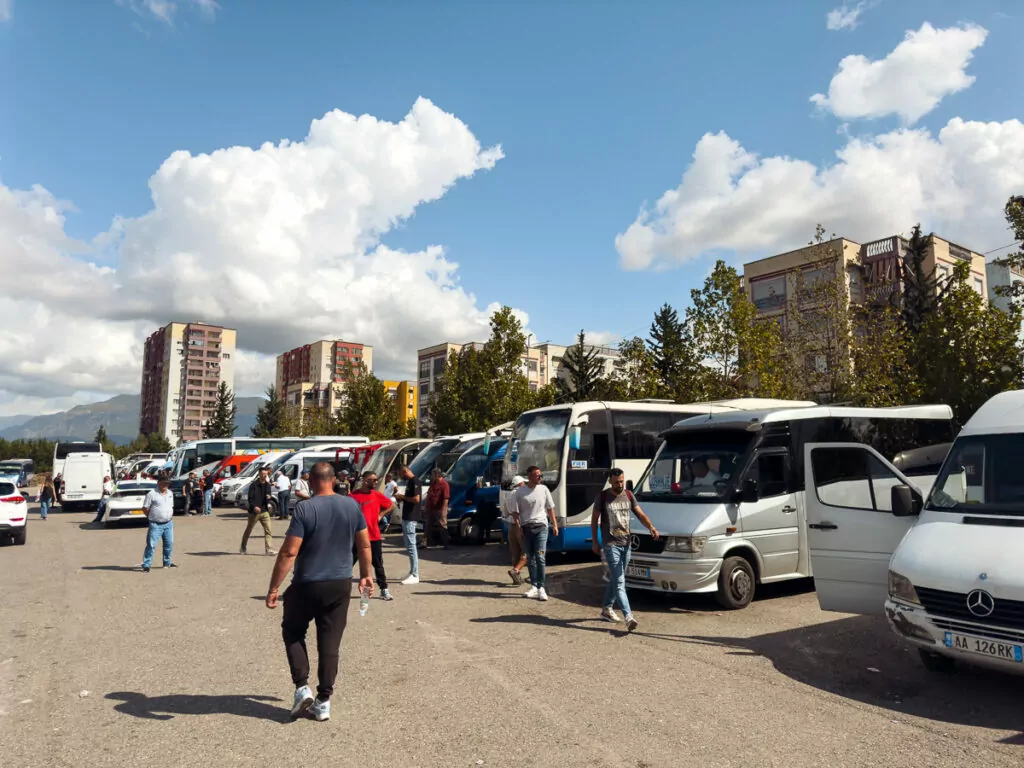 Bus station in Tirana shows Albania solo travel infrastructure, with tourists and locals navigating between parked coaches under mountain views.