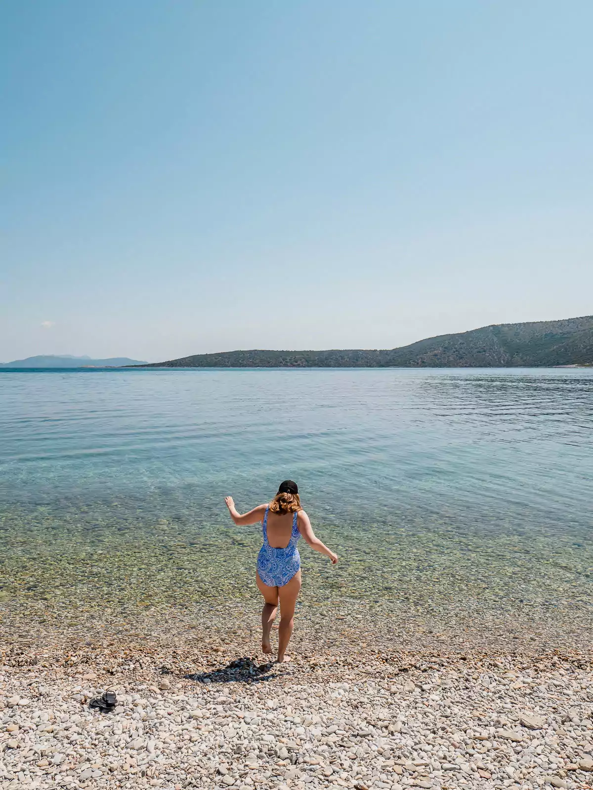 A woman wading into the tranquil, crystal-clear sea from a pebble beach under a bright blue sky. What to know before going to Greece? Its beaches are perfect for both serenity and adventure