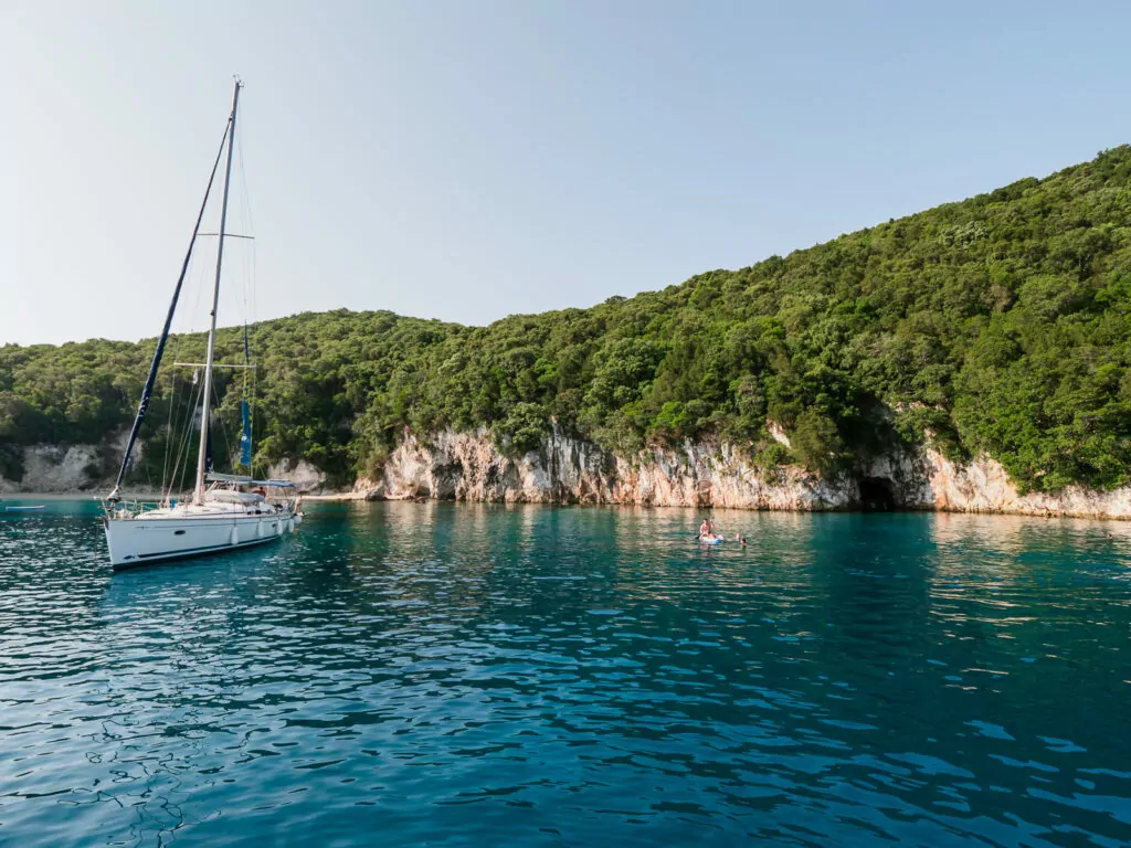 A white sailboat anchored in a quiet cove, surrounded by lush green cliffs and clear blue water. Greek islands travel tips often highlight secluded spots like this for relaxation and exploration