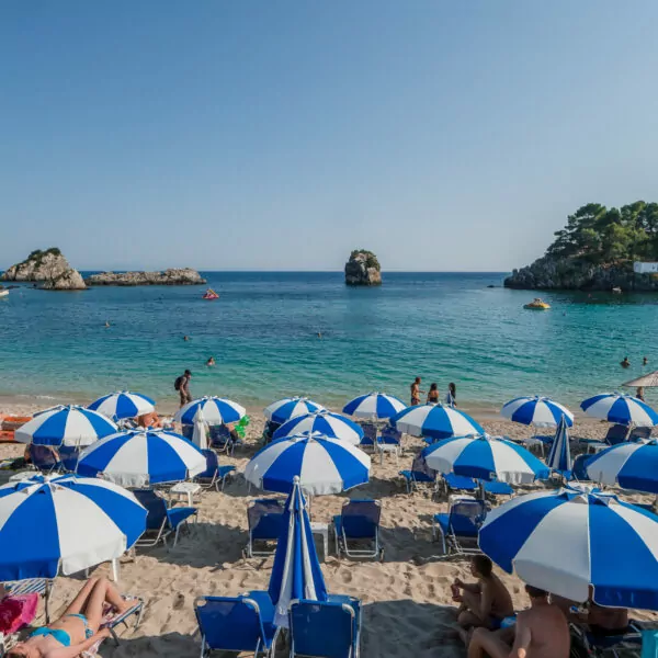 A vibrant beach in Parga, Greece, lined with blue-and-white striped umbrellas and sun loungers, overlooking the clear waters of the Greek islands