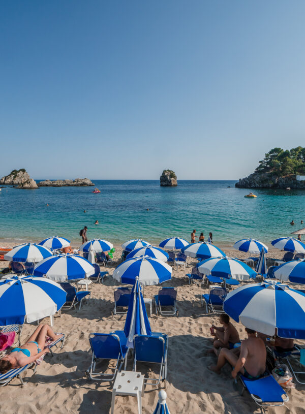 A vibrant beach in Parga, Greece, lined with blue-and-white striped umbrellas and sun loungers, overlooking the clear waters of the Greek islands