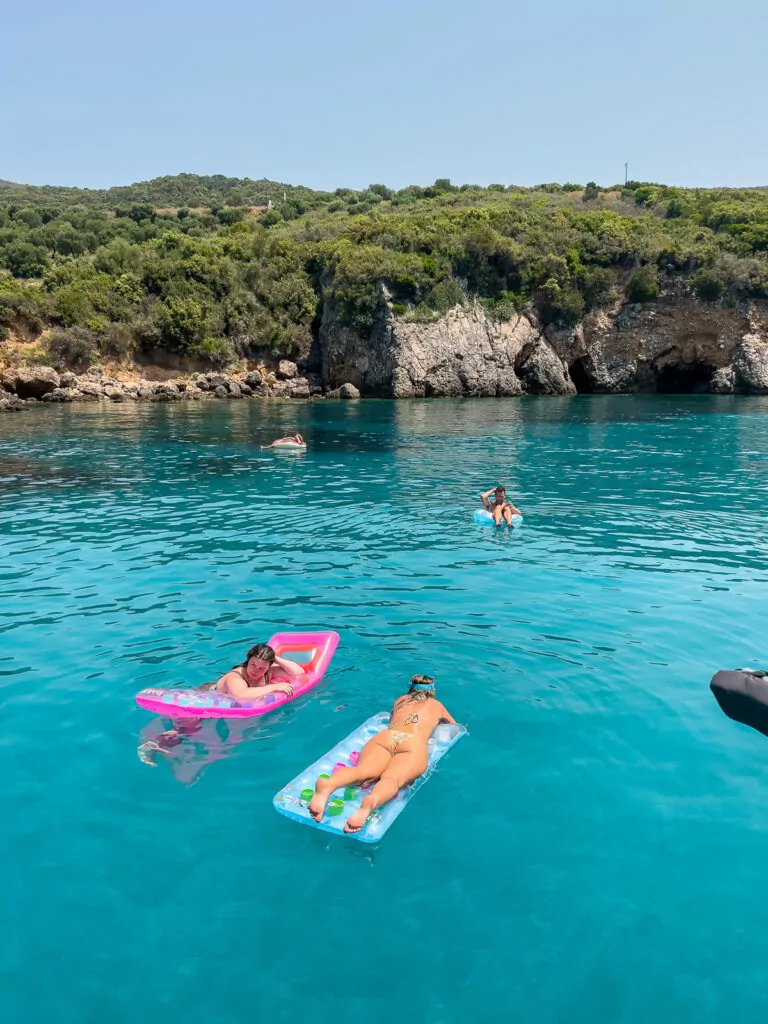 Visitors floating on colourful inflatables in the crystal-clear waters near the rocky coast of Corfu, Greece. Essential tips for travelling to Greece include exploring hidden swimming spots like this