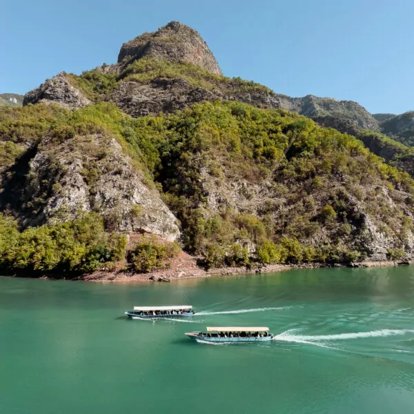 Tour boats create white wakes on Lake Koman's green waters as they ferry visitors between towering cliffs