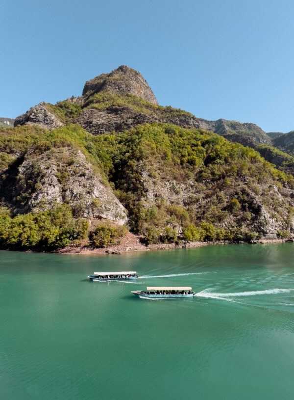 Tour boats create white wakes on Lake Koman's green waters as they ferry visitors between towering cliffs