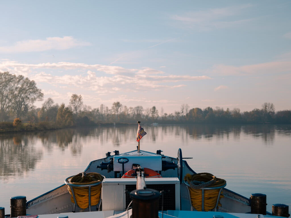 The bow of the barge gliding through calm waters, with the French flag flying and the Burgundian countryside reflected in the morning light.