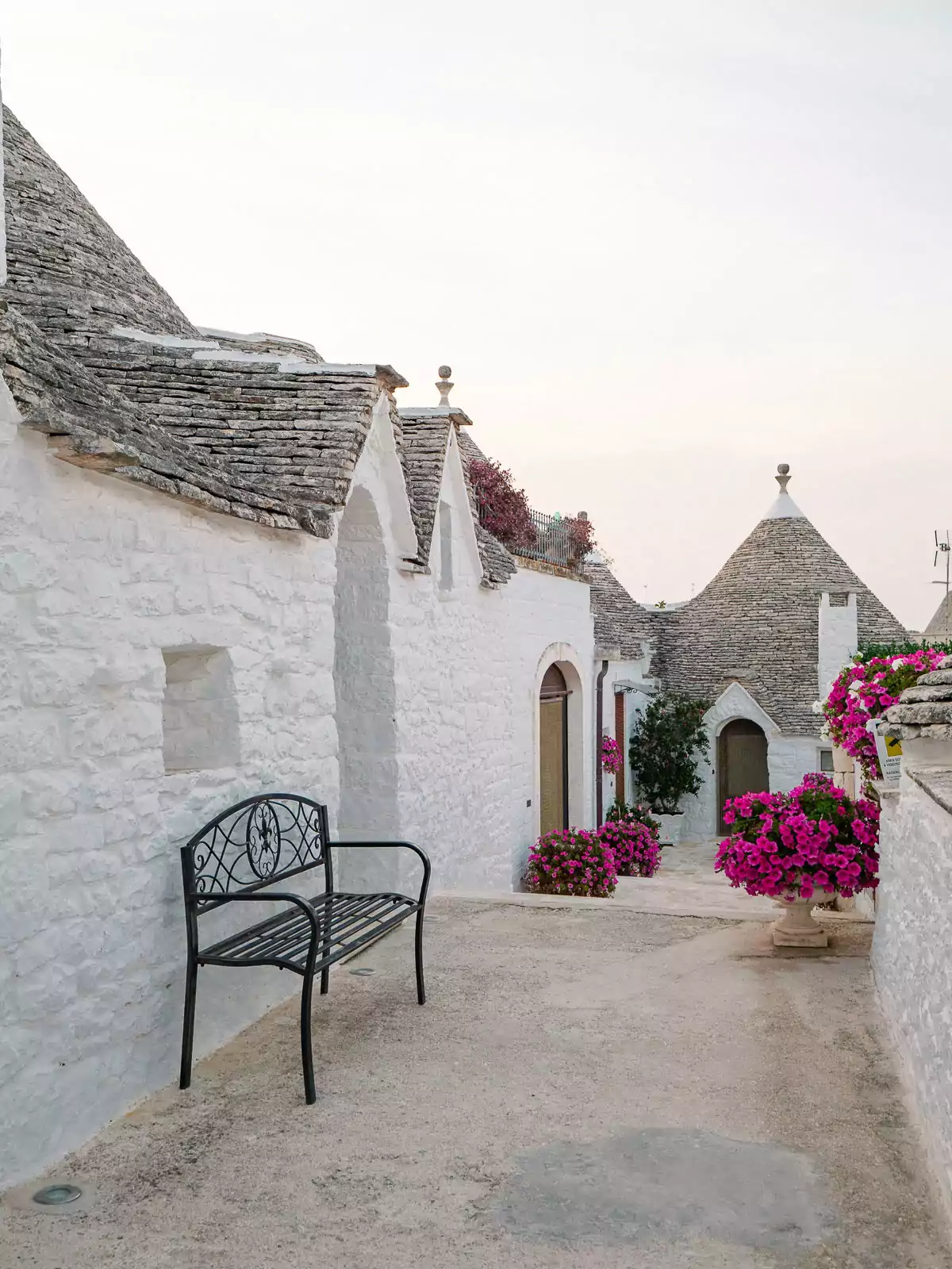 A peaceful alleyway between traditional trulli houses in Alberobello, with a wrought iron bench and bright pink bougainvillea flowers, demonstrating how Puglia without a car allows for intimate exploration of these UNESCO sites.
