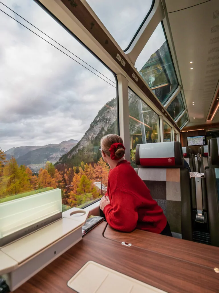 Woman in red gazing at autumn scenery from inside the Glacier Express