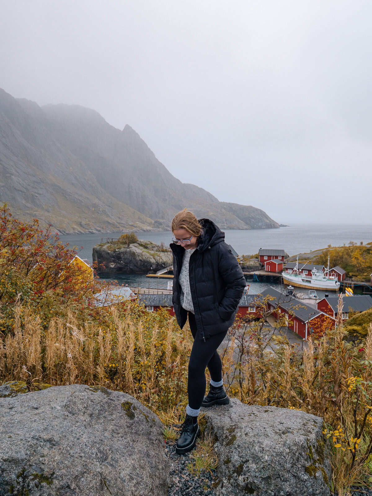 Woman standing on a rocky outcrop in Lofoten, overlooking a small fishing village with colourful houses nestled between the sea and steep coastal mountains. A perfect starting point for a two weeks in Norway adventure
